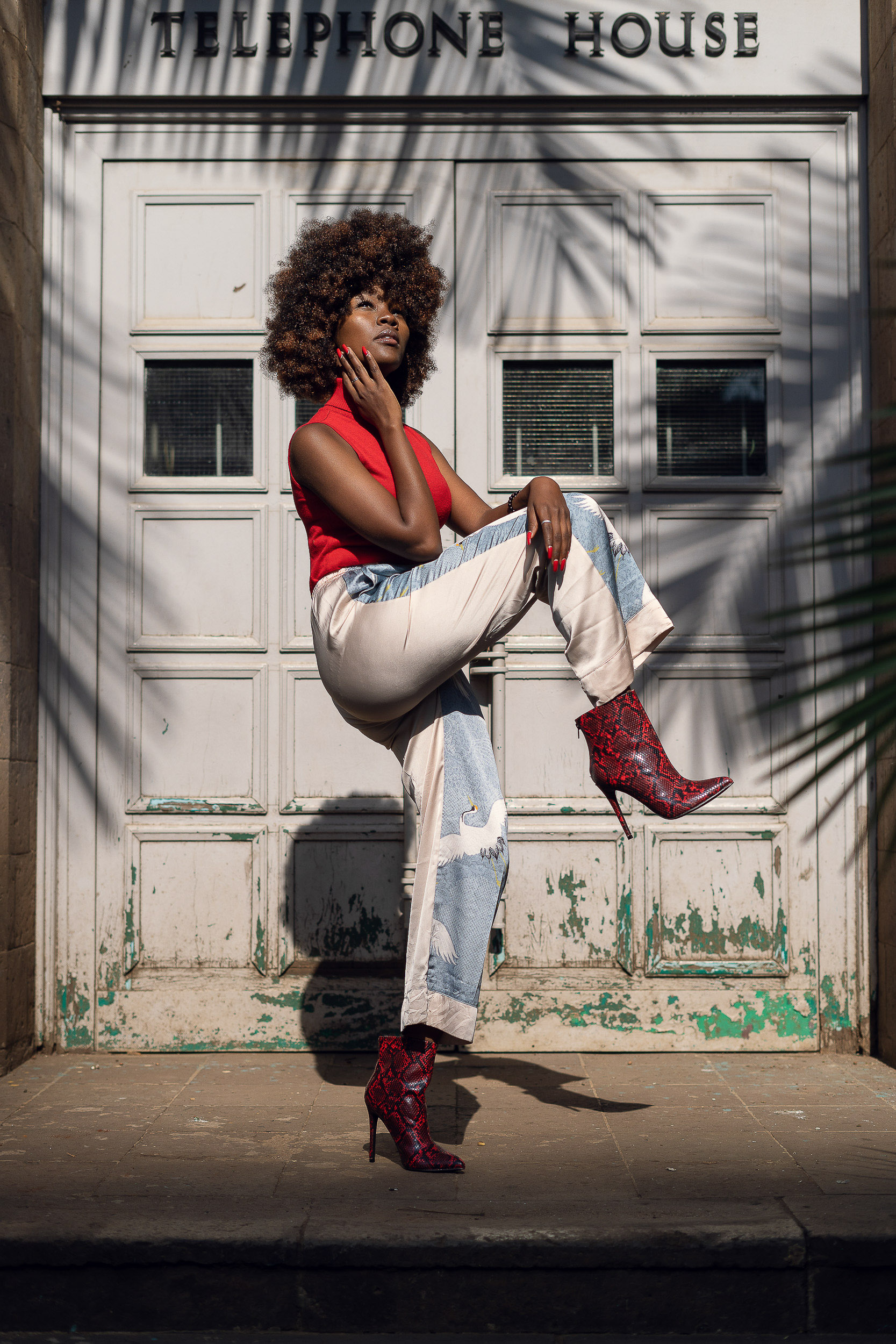 Street fashion portrait of a woman in a red top, wide jeans and red boots outside a building marked Telephone House