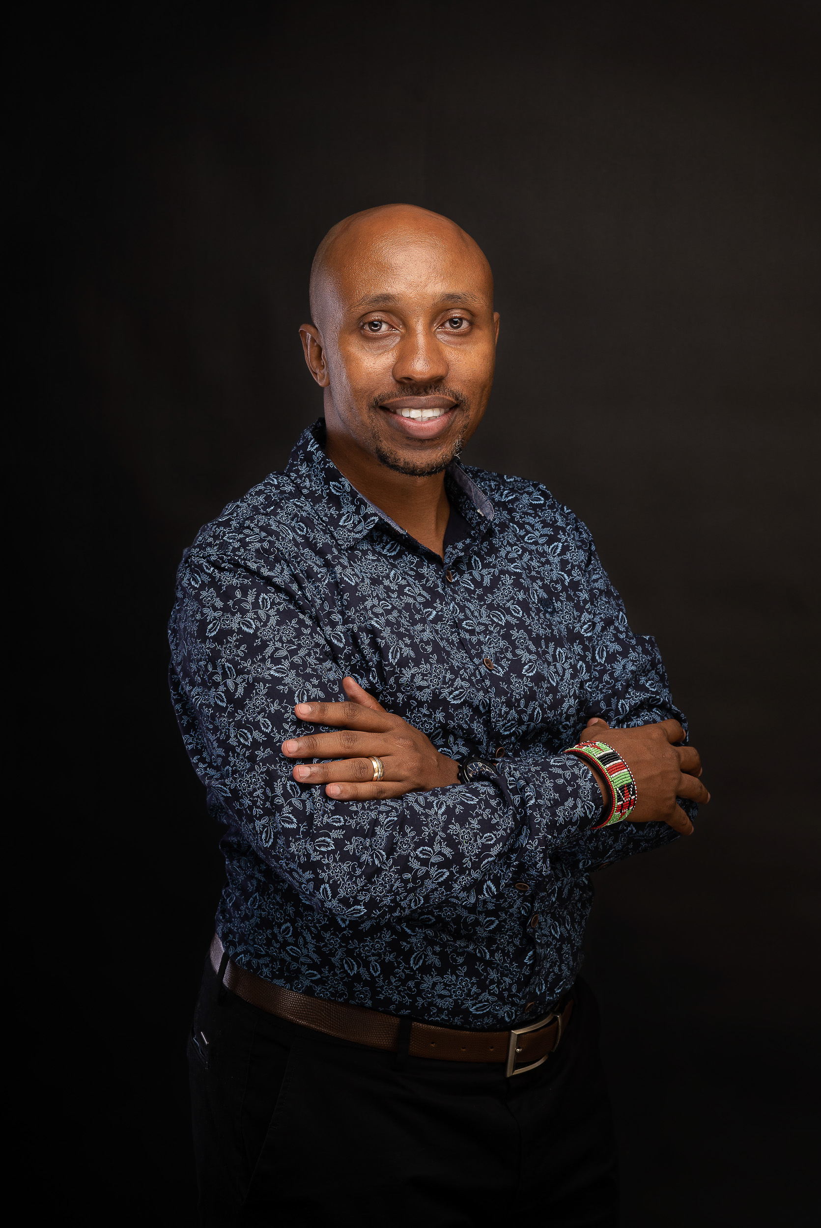 Corporate headshot of a smiling man in a patterned shirt with arms folded on a black background